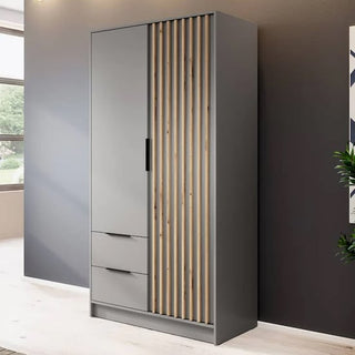The CHOICEWARDROBE Newberry 105cm Grey 2 Door Wardrobe, featuring an oak slatted design, drawers, and shelves, stands against a dark wall in a bright room with a window and nearby plant.