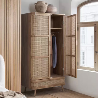 The CHOICEWARDROBE Nixa Oak Rattan Wooden Wardrobe (90 cm, 2 doors, 1 drawer) with cane panels stands open, displaying hanging space and a drawer. Two ceramic vases sit on top by a window and wood-paneled wall in a bright room.