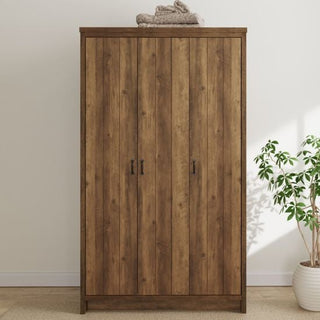The CHOICEWARDROBE Nolan Knotty Oak 3 Door Wardrobe, featuring a rustic wooden design with black handles, stands by a white wall. A folded towel is on top, and a green potted plant rests nearby on the light floor.