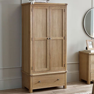 The CHOICEWARDROBE Norland Limed Oak 2 Door Wardrobe, featuring a bottom drawer and classic farmhouse style, stands on a wooden floor beside a beige wall. A circular mirror and small decor items sit on an adjacent cabinet.