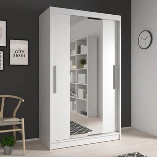 The CHOICEWARDROBE Novaro 2 Door Sliding Wardrobe in gloss white stands against a dark wall, reflecting a bookshelf. A wooden chair and framed artwork are placed on the left, enhancing the modern bedroom storage's fresh look.