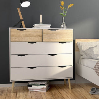 The CHOICEWARDROBE Okla 3 Piece Bedroom Furniture Set in white & oak features Scandinavian minimalist style—a 4 drawer chest topped with a lamp, candle, and vase of yellow flowers, books below, and a bed with a white pillow partially visible nearby.
