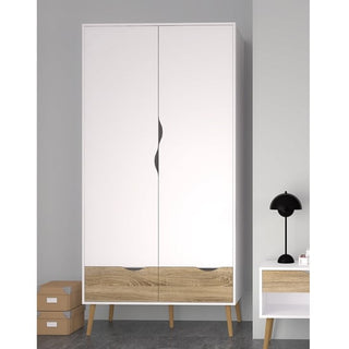 The CHOICEWARDROBE Oklo Scandinavian Wardrobe, featuring 2 white doors, 2 oak drawers, and tapered legs, stands beside a small white nightstand with an oak drawer, a black lamp, and two stacked cardboard boxes.
