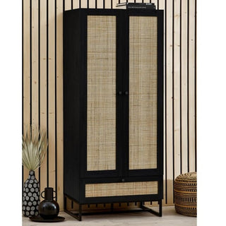 The CHOICEWARDROBE Padstow Black Rattan Wardrobe – 2 Door + 1 Drawer (80cm) stands against a wood-paneled wall, styled with a black vase and patterned pouf—perfect boho modern bedroom storage or a chic wardrobe.