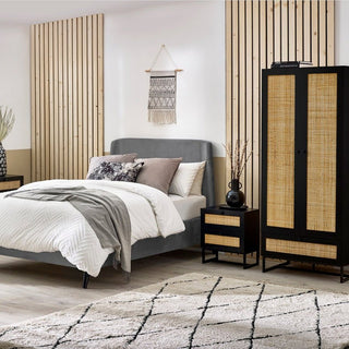 A modern bedroom features a gray upholstered bed, white bedding, and neutral pillows. The CHOICEWARDROBE Padstow Black Rattan Wardrobe adds boho-modern storage, paired with a matching nightstand, wood slats, and a geometric rug for a stylish finish.