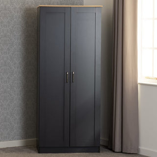 The CHOICEWARDROBE Parnu 2 Door Panelled Wooden Wardrobe in charcoal grey with an oak effect top and silver handles stands against patterned wallpaper near a window with beige curtains in a carpeted room.