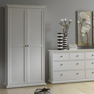 The CHOICEWARDROBE Paroya 96cm 2 Door Wooden Wardrobe in white with paneled design stands by a gray wall, paired with a matching dresser. Vases, books, and floral art add elegance, while the light-tiled floor completes the serene bedroom look.
