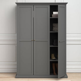 The CHOICEWARDROBE Paroya Wooden Wardrobe in classic grey with 3 doors, paneled detailing, and brass knob handles stands against a white-paneled wall. One door is ajar, revealing shelves with books, a folded blanket, shoes, and partially visible human legs.