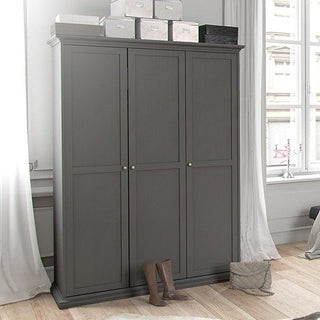 The CHOICEWARDROBE Paroya Wooden Wardrobe, 3 doors and classic grey finish with paneled detailing and brass knob handles, stands against a white wall by a window. Boxes are stacked atop, while boots and a quilted bag rest on the floor in front.