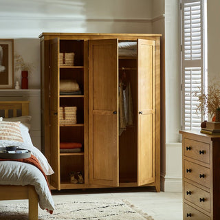 The CHOICEWARDROBE Porsha 142cm 3 Door Waxed Oak Wardrobe, featuring a solid wooden frame, open doors to show shelves with folded clothes and a hanging rail for garments beside a bed, window, and chest of drawers in a sunlit bedroom.
