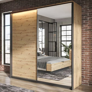 The CHOICEWARDROBE Qesso 220cm 2 Door Sliding Mirrored Wardrobe in oak finish with a black frame and LED lighting stands by a brick wall, reflecting a modern bedroom with a bed, large windows, plant, and glass doors.