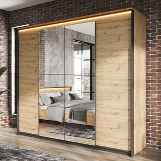 The CHOICEWARDROBE Qesso 4 Door Hinged Mirrored Wardrobe in 250cm Rustic Oak with LED Light and Black Frame stands against a brick wall, reflecting a modern bedroom and bringing stylish functionality to the space.