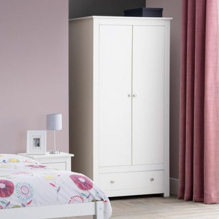 The CHOICEWARDROBE Raddix 2 Door Wardrobe with Drawer in Surf White stands in a pink bedroom corner. Next to it, a white bedside table holds a lamp and photo frame, with part of a bed displaying floral bedding visible.