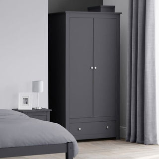 The CHOICEWARDROBE Radley 2 Door Wardrobe in Anthracite Grey, featuring a compact 97cm design with a hanging rail and lower drawer, stands in the bedroom corner, complemented by a bed, nightstand, lamp, framed photo, grey curtains, and light flooring.
