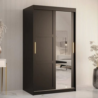 The CHOICEWARDROBE Rieti Wardrobe 100cm in black features 2 sliding doors—one full-length mirrored and one plain, both with gold handles—standing against a striped wall and reflecting a bed and black bedroom furniture.
