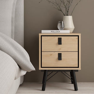 A CHOICEWARDROBE Rynok Oak and Black bedside table with metal legs from the 3 Piece Bedroom Furniture Set stands by the bed, topped with a beige vase, glass, and "Pollock," while a taupe wall adds warmth to the industrial bedroom.