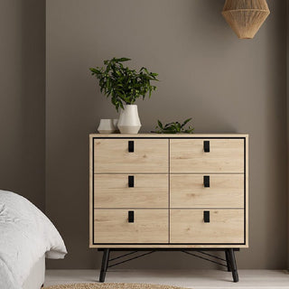 An oak and black CHOICEWARDROBE Rynok 3 Piece Bedroom Set with metal legs stands by a taupe wall. The set includes a 3 door wardrobe, 4 drawer chest, and bedside table, accented by white vases, a plant, and nearby white bedding.
