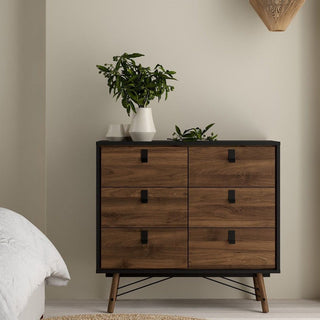 The CHOICEWARDROBE Rynok 3 Piece Wooden Bedroom Furniture Set in walnut & black finish, featuring a chest of drawers, is set against a beige wall with white vases and leafy plants near a white bed and woven rug.