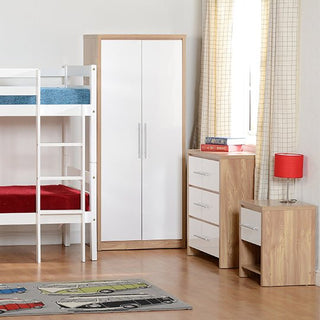 A children’s bedroom features a white bunk bed, the CHOICEWARDROBE Samaira White Gloss 2 Door Wardrobe with Oak Effect Frame, a chest of drawers with a red lamp and books, and sunlight spilling over checkered curtains onto a car-themed rug.