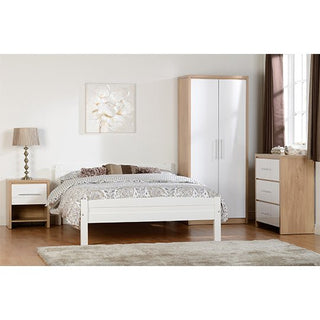A bedroom with a white bed, patterned bedding, wooden nightstand and lamp, CHOICEWARDROBE Samaira White Gloss 2 Door Wardrobe with Oak Effect Frame, matching dresser, beige rug, large window with brown curtains, and a floral painting above the bed.