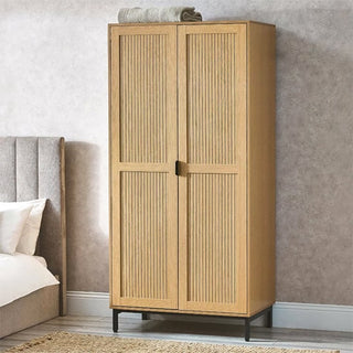 The CHOICEWARDROBE Sarnia Natural Oak 2 Door Wardrobe, featuring slatted wood doors and metal legs in a Japandi modern design, stands by a bed against a light wall with a folded grey towel placed on top.