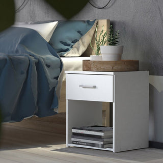 A CHOICEWARDROBE Scalia white bedside table from the 3 Piece Bedroom Furniture Set, featuring a drawer and open shelf with books, stands by a bed with blue bedding and two potted plants—ideal for modern bedrooms.