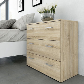 A CHOICEWARDROBE Scalia Oak 3 Drawer Chest, part of the 3 Piece Wooden Bedroom Set in natural light oak with silver handles, stands on a gray floor next to a bed. A glass vase with white flowers sits on top and art hangs above on the white wall.