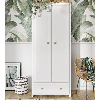 The CHOICEWARDROBE Sevilla I Wooden 2 Door Wardrobe in Matt White & Oak (85cm) with drawer and hanging rail stands against green leaf-patterned wallpaper, paired with a rattan chair, striped basket, framed art, and greenery above.