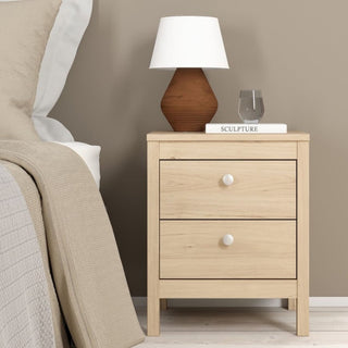 A light oak-effect bedside table with two drawers from the CHOICEWARDROBE Solara 3-Piece Bedroom Furniture Set sits by the bed, topped with a brown and white lamp, a glass of water, and a book. Neutral bedding and walls complete the look.
