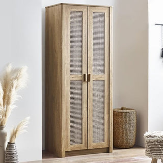 The CHOICEWARDROBE Sumter Oak & Rattan 2 Door Wardrobe, featuring a natural wooden frame and woven cane panels in a boho modern design, stands by a white wall beside a woven basket and pampas grass in a vase.