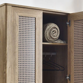 The CHOICEWARDROBE Sumter Oak & Rattan 2 Door Wardrobe, featuring a natural wooden frame and woven cane panels, is partially open to reveal a rolled blanket and empty black hangers—ideal for a boho-modern oak bedroom collection.