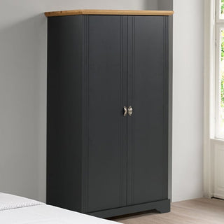 The CHOICEWARDROBE Talox 100cm Grey Painted Wooden 2 Door Wardrobe with Oak Top stands against a light gray wall by a window, its silver handles visible. Part of a bed with a white blanket appears in the foreground.