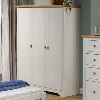 The CHOICEWARDROBE Talox 142cm White Wooden 3 Door Wardrobe with Oak Top stands against a light green wall. Silver handles, a plant, and a basket accent it, with a matching chest of drawers partly visible on the right.