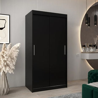 The CHOICEWARDROBE Tavira 100cm Black 2 Door Sliding Wardrobe, with a modern matte finish and sleek silver handles, complements a contemporary bedroom with a round mirror, makeup table, abstract art, and vertical wall paneling.