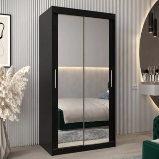 The CHOICEWARDROBE Tavira 100cm Black Mirrored Sliding Door Wardrobe, with a compact modern design and storage shelves, is featured in a stylish bedroom with a green-blanketed bed, vertical wall paneling, pampas grass, and a round mirror.