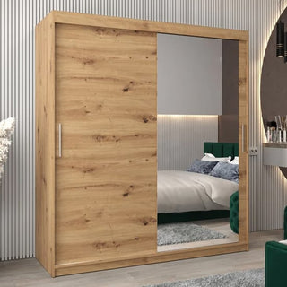 The CHOICEWARDROBE Tavira 2 Door Oak Effect Sliding Wardrobe (180cm) with shelves and hanging rail stands in a modern bedroom, alongside a bed, green upholstered furniture, and a circular wall mirror.