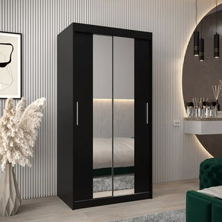 The CHOICEWARDROBE Tavira 2 Door Sliding Mirrored Wardrobe in black stands in a modern bedroom with a green sofa, round wall mirror, plant, and pendant lights against a vertically grooved wall.