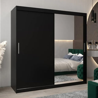 The CHOICEWARDROBE Tavira 2 Door Sliding Mirrored Wardrobe in black matte finish stands in a modern bedroom with a green upholstered bed, gray walls, and a small dressing table on the right.
