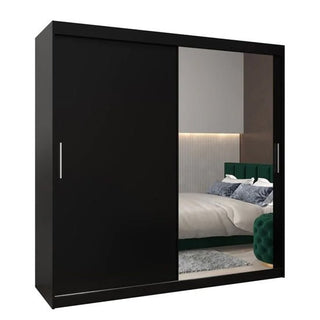 The CHOICEWARDROBE Tavira 2 Door Sliding Mirrored Wardrobe in black matte adds modern style, reflecting a bedroom with a green upholstered headboard, white and blue bedding, and a textured accent wall.