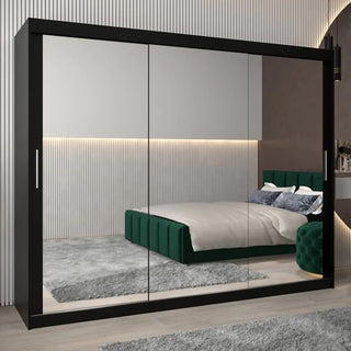 The CHOICEWARDROBE Tavira 3 Door Mirrored Sliding Wardrobe in black adds modern style, with full mirror panels reflecting a green upholstered bed, grey rug, and wall accent lighting in the bedroom.