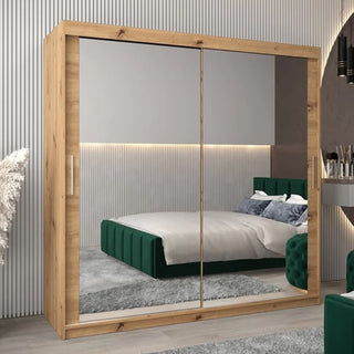 The CHOICEWARDROBE Tavira 3 Mirror Sliding Door Wardrobe (203cm, Oak Effect) enhances a modern bedroom with its mirrored doors reflecting a green bed, gray rug, striped walls, and soft lighting. Features shelves and hanging rail for storage.