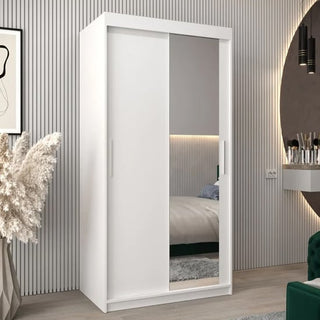 The CHOICEWARDROBE Tavira II 100cm Mirrored Sliding Door Wardrobe in white, with shelves and drawers, stands in a modern bedroom with a striped wall and decorative grass, reflecting the bed and offering stylish, compact storage.