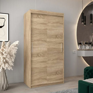 The CHOICEWARDROBE Tavira Wardrobe 100cm in Sonoma Oak with 2 sliding doors, hanging rail, and shelving stands on a light floor beside a modern vanity, round mirror, green sofa, and pampas grass in this contemporary bedroom.