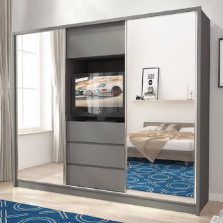 A modern bedroom featuring the ChoiceWardrobe Telluride Mirrored Sliding Door Wardrobe in Graphite (254cm), with central drawers and TV space, beige bedding on the bed, white walls, and a blue patterned carpet.