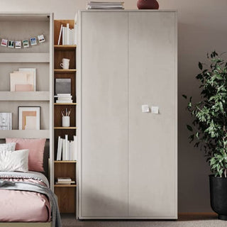 A neatly made bed with pink and white bedding sits beside a bookshelf and the CHOICEWARDROBE Tifton Silk Finish Wooden Wardrobe—101 cm, 2-door design—while books, decor, and a potted plant complete the cozy setting.