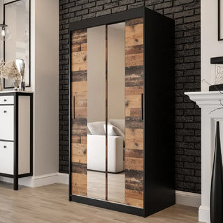 The CHOICEWARDROBE Topeka 100cm Dark Oak & Black Sliding Mirrored Wardrobe in a modern industrial style stands on a light floor by a black brick wall, beside white furniture and a fireplace in a contemporary bedroom.