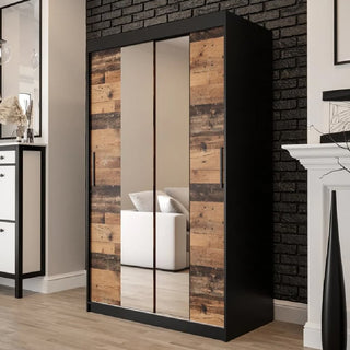The CHOICEWARDROBE Topeka 120cm Dark Oak & Black Sliding Wardrobe with 2 Mirror Doors stands against a black brick wall, its mirrored panels reflecting a white couch in this modern industrial bedroom.