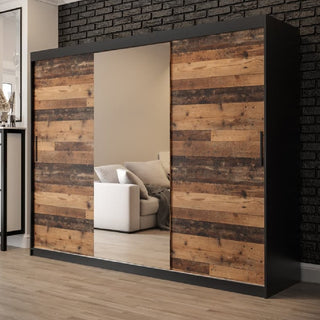 The CHOICEWARDROBE Topeka 3 Door Mirrored Sliding Wardrobe in Rustic Oak & Black stands against a black brick wall in a modern room with wood flooring, its full-length mirror reflecting a white couch with cushions.