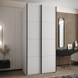 A modern bedroom showcases the CHOICEWARDROBE Tressaro 100cm Sliding 2 Door Wardrobe in matte white with black trim, set against paneled and wooden accent walls. Nearby, a black desk with a mirror and chair offers extra storage and style.