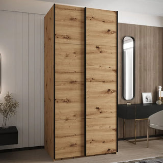 The CHOICEWARDROBE Tressaro Artisan Oak Sliding Wardrobe – 2 Door (100cm, Rustic Grain Finish) with black accents stands in a modern bedroom with contrasting walls, a small desk, mirror, and décor for practical storage.